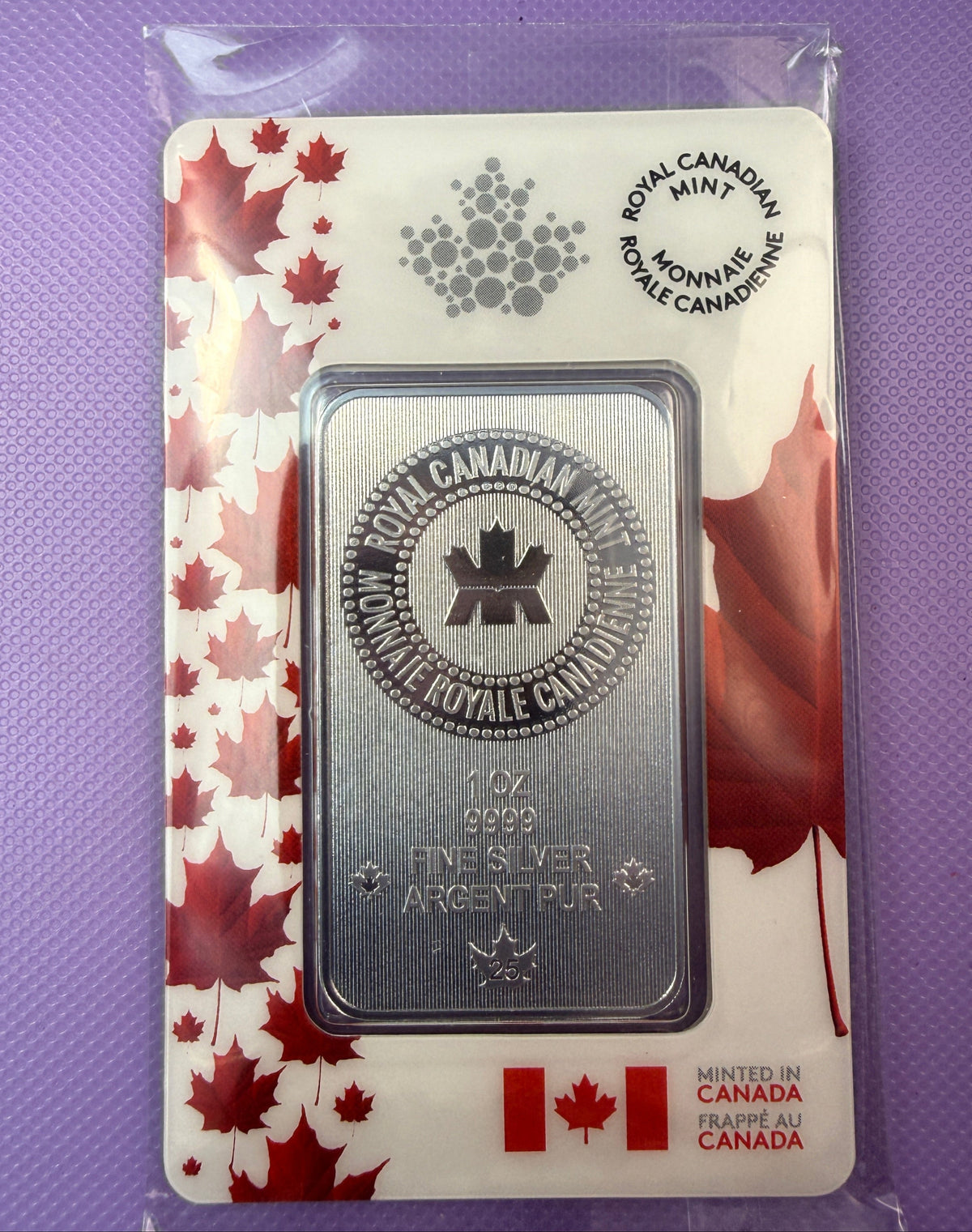 A 1 oz silver bullion bar from the Royal Canadian Mint, presented in its original plastic packaging with a white and red maple leaf card backing, set against a purple textured background.